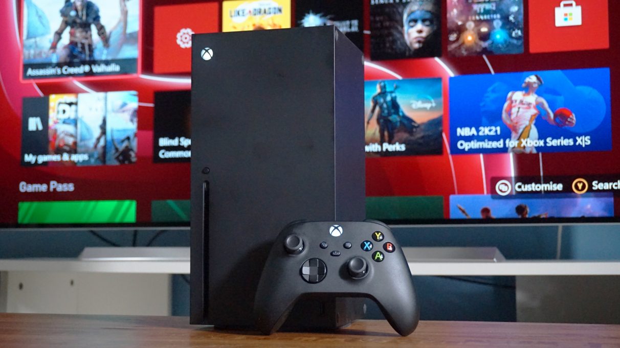 A photo of the Xbox Series X standing upright with its controller in front of a TV.